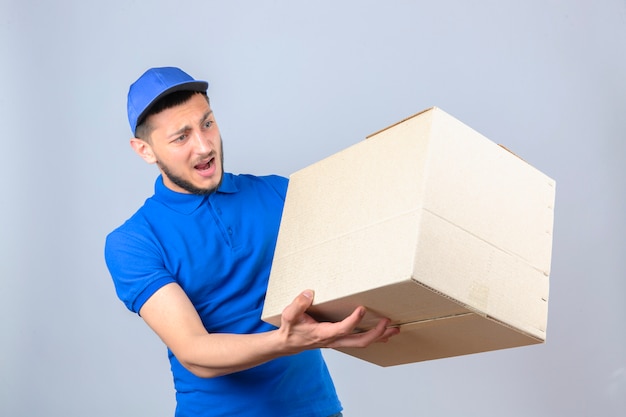 Jovem entregador vestindo camisa pólo azul e boné parecendo surpreso e confuso em pé com uma caixa de papelão sobre um fundo branco isolado