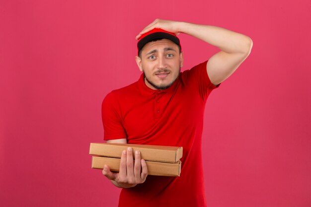 Foto grátis jovem entregador vestindo camisa pólo vermelha e boné em pé com caixas de pizza surpreso com a mão na cabeça por engano, lembre-se de erro esqueceu o conceito de memória ruim sobre fundo rosa isolado