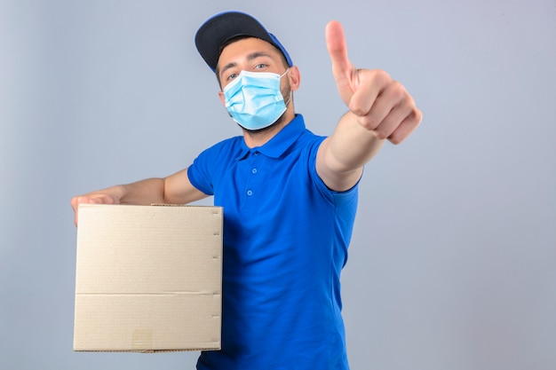 Jovem entregador vestindo uma camisa pólo azul e boné com máscara protetora médica em pé com uma caixa de papelão mostrando o polegar para a câmera sobre fundo branco isolado