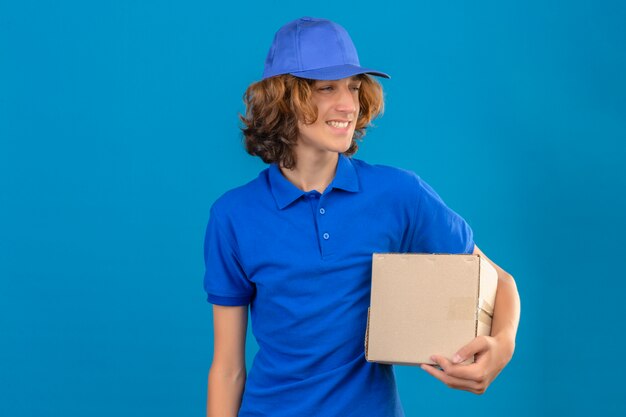 Jovem entregador vestindo uma camisa pólo azul e boné segurando uma caixa de papelão sorrindo amigável em pé sobre fundo azul