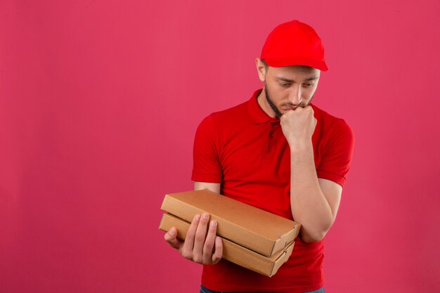 Jovem entregador vestindo uma camisa pólo vermelha e boné em pé com uma pilha de caixas de pizza, parecendo triste e infeliz olhando de lado com a mão pressionada contra o queixo sobre um fundo rosa isolado