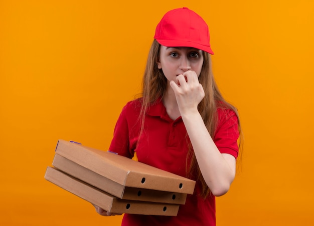 Jovem entregadora ansiosa de uniforme vermelho segurando pacotes com a mão nos lábios em um espaço laranja isolado