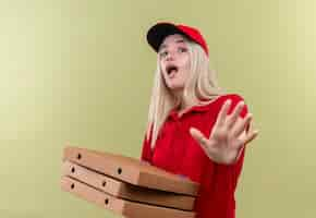 Foto grátis jovem entregadora assustada com uma camiseta vermelha com boné segurando uma caixa de pizza e mostrando um gesto de parada