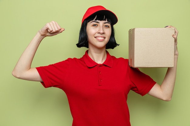 Jovem entregadora caucasiana sorridente segurando uma caixa de papelão e tensionando os bíceps