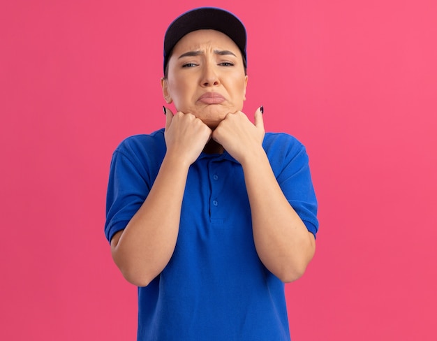 Jovem entregadora chateada com uniforme azul e boné olhando para a frente com uma expressão triste franzindo os lábios em pé sobre a parede rosa