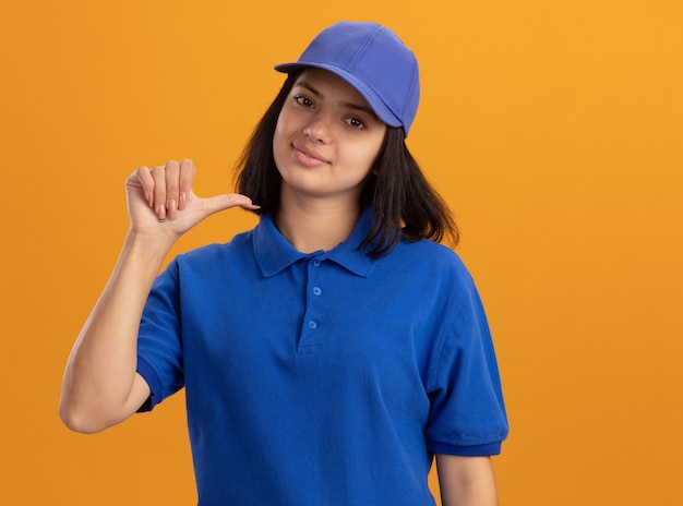 Jovem entregadora com uniforme azul e chapéu apontando para si mesma, sorrindo confiante em pé sobre a parede laranja