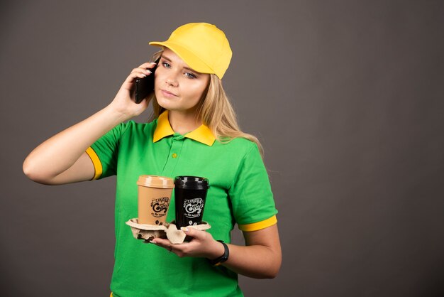 Jovem entregadora com xícaras de café, falando no smartphone.