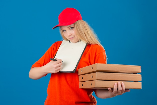 Jovem entregadora de camisa pólo laranja e boné vermelho com máscara de proteção médica em pé com uma pilha de caixas de pizza e prancheta com caneta pedindo assinatura sobre background azul isolado