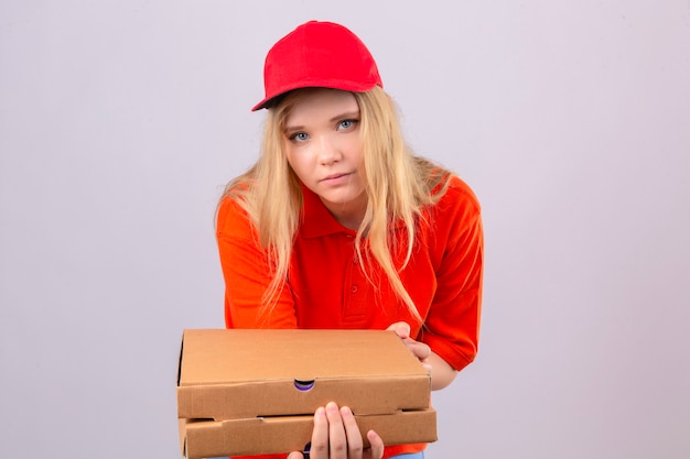 Jovem entregadora de camisa pólo laranja e boné vermelho, olhando para a câmera estendendo uma pilha de caixas de pizza sobre um fundo branco isolado