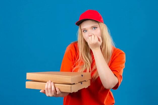 Jovem entregadora de camisa pólo laranja e boné vermelho segurando uma pilha de caixas de pizza, parecendo estressada e nervosa, com as mãos na boca roendo as unhas sobre um fundo azul isolado