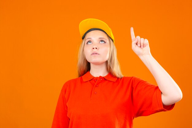 Jovem entregadora de camisa pólo vermelha e boné amarelo, olhando para cima e apontando com o dedo indicador sobre um fundo laranja isolado