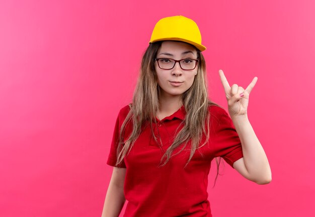 Jovem entregadora de camisa pólo vermelha e boné amarelo, parecendo confiante fazendo o símbolo do rock com finfers