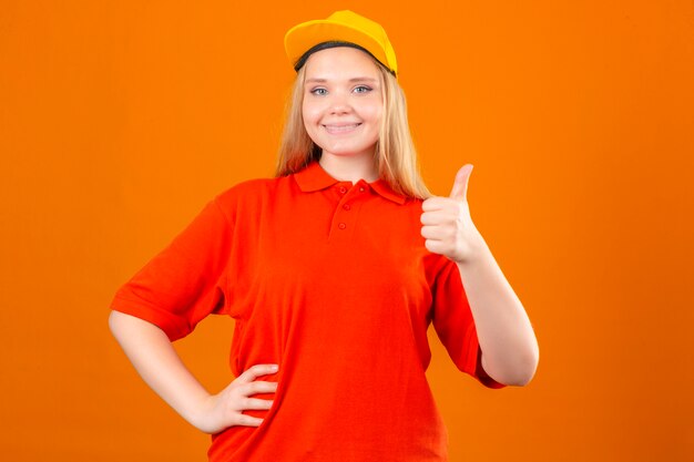 Jovem entregadora de camisa pólo vermelha e boné amarelo, parecendo confiante, mostrando o polegar sobre um fundo laranja isolado