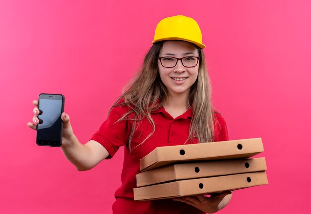 Jovem entregadora de camisa pólo vermelha e boné amarelo segurando uma pilha de caixas de pizza, olhando para a câmera com um sorriso confiante no rosto, mostrando o smartphone