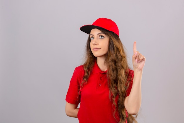 Jovem entregadora de camisa pólo vermelha e boné apontando o dedo pensando sobre um fundo branco isolado