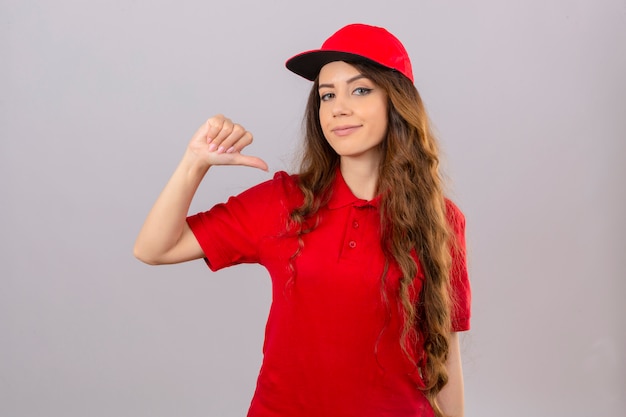 Jovem entregadora de camisa pólo vermelha e boné apontando o polegar para si mesma, sorrindo amigavelmente sobre um fundo branco isolado