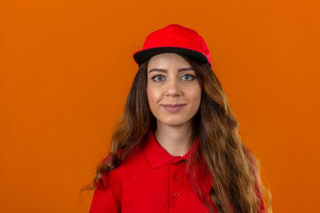 Jovem entregadora de camisa pólo vermelha e boné com cabelo encaracolado, olhando para a câmera com um sorriso confiante sobre fundo laranja isolado