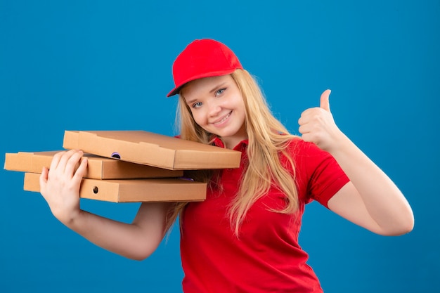 Jovem entregadora de camisa pólo vermelha e boné em pé com caixas de pizza, parecendo confiante, sorrindo alegremente mostrando o polegar para cima sobre um fundo azul isolado