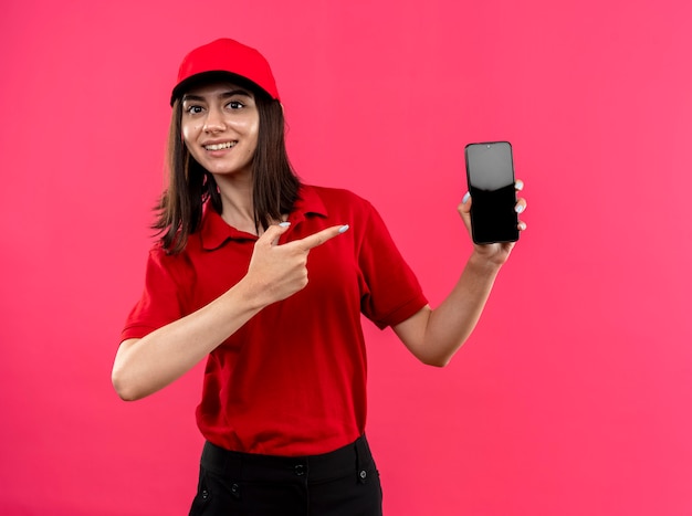 Jovem entregadora de camisa pólo vermelha e boné mostrando o smartphone apontando com o indicador de fionger para ele sorrindo alegremente em pé sobre a parede rosa