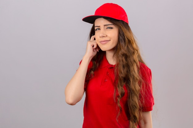 Jovem entregadora de camisa pólo vermelha e boné, olhando para a câmera, olhando para a câmera com os olhos semicerrados, suspeitando de um fundo branco isolado