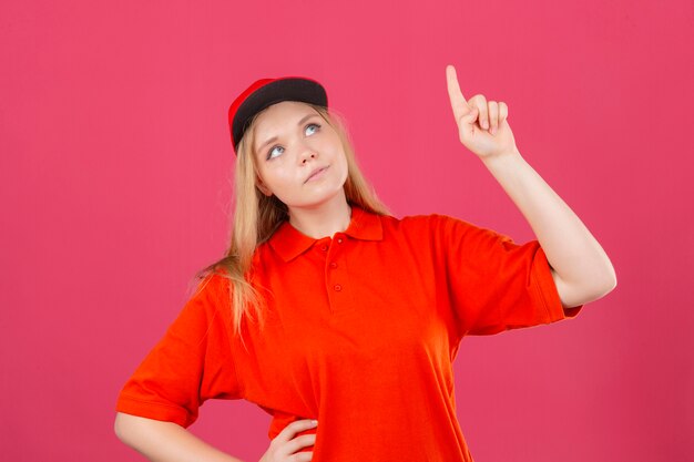 Jovem entregadora de camisa pólo vermelha e boné olhando para cima e apontando para algo com o dedo indicador sobre fundo rosa isolado