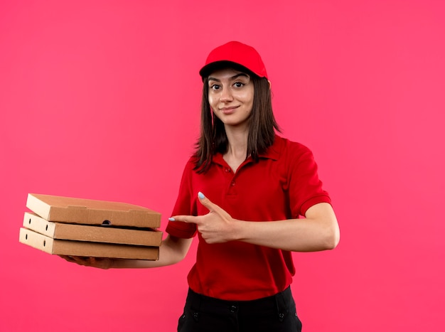 Jovem entregadora de camisa pólo vermelha e boné segurando caixas de pizza apontando com o dedo indicador para elas, sorrindo amigavelmente em pé sobre a parede rosa
