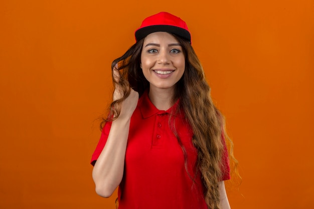 Jovem entregadora de camisa pólo vermelha e boné torcendo uma mecha de cabelo no dedo, sorrindo alegremente sobre um fundo laranja isolado