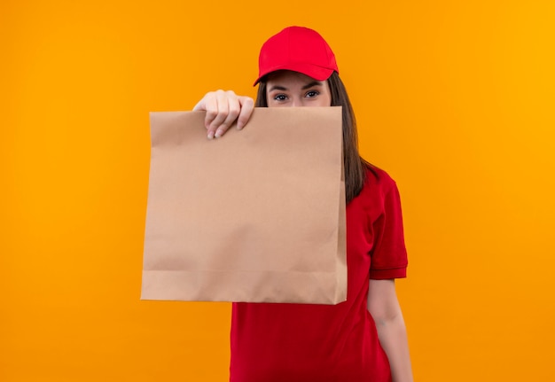 Foto grátis jovem entregadora de camiseta vermelha com boné vermelho segurando um pacote sobre fundo amarelo