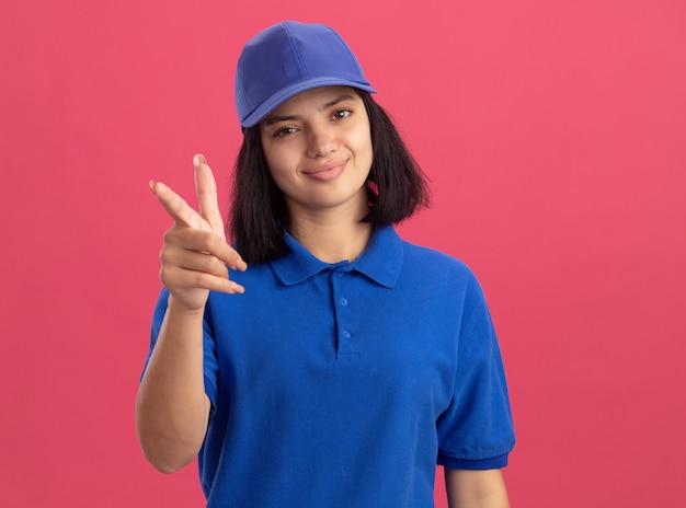 Jovem entregadora de uniforme azul e boné apontando com o dedo indicador sorrindo amigavelmente em pé sobre a parede rosa