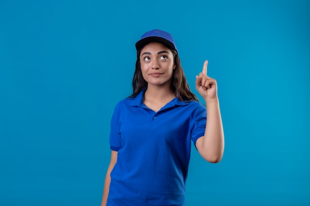 Jovem entregadora de uniforme azul e boné apontando o dedo para cima, concentrada em uma tarefa em pé sobre um fundo azul