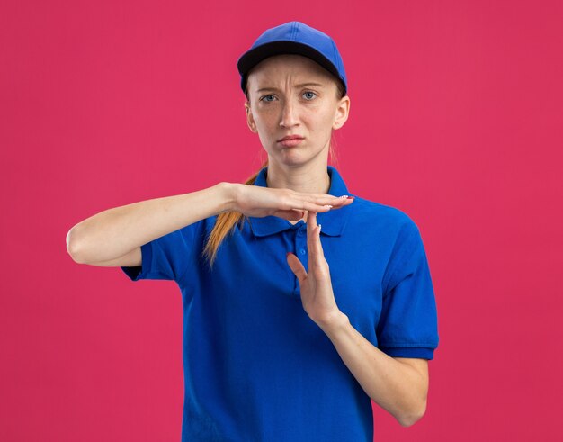 Jovem entregadora de uniforme azul e boné com cara séria fazendo gesto de castigo com as mãos em pé sobre a parede rosa