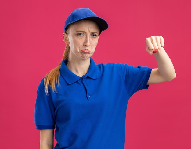 Jovem entregadora de uniforme azul e boné com expressão triste franzindo os lábios e mostrando o punho