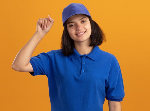 Jovem entregadora de uniforme azul e boné com o punho cerrado, sorrindo em pé sobre a parede laranja