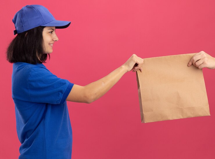 Foto grátis jovem entregadora de uniforme azul e boné dando um pacote de papel para um cliente sorrindo amigável em pé sobre uma parede rosa