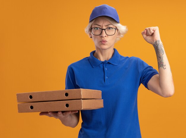 Jovem entregadora de uniforme azul e boné de óculos segurando caixas de pizza com cara séria levantando o punho sobre a parede laranja