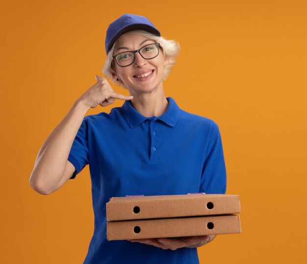 Jovem entregadora de uniforme azul e boné de óculos segurando caixas de pizza, sorrindo, fazendo um gesto para mim, sorrindo por cima da parede laranja