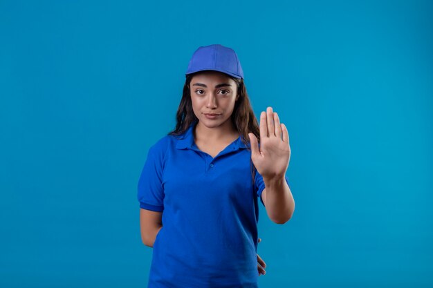 Jovem entregadora de uniforme azul e boné em pé com a mão aberta, fazendo sinal de pare com gesto de defesa de expressão sério e confiante sobre fundo azul