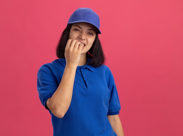 Jovem entregadora de uniforme azul e boné estressada e nervosa roendo as unhas em pé sobre uma parede rosa