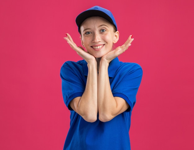 Jovem entregadora de uniforme azul e boné feliz e positiva, sorrindo alegremente com a mão no rosto