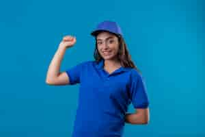 Foto grátis jovem entregadora de uniforme azul e boné levantando o punho, olhando para a câmera, sorrindo confiante e alegre por seu sucesso e vitória em pé sobre um fundo azul