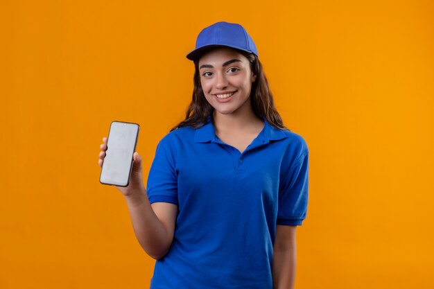 Jovem entregadora de uniforme azul e boné mostrando smartphone sorrindo alegremente em pé sobre fundo amarelo