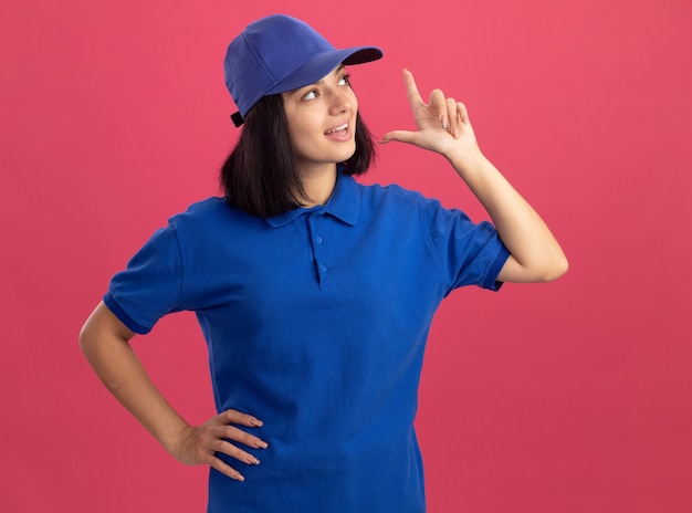 Foto grátis jovem entregadora de uniforme azul e boné olhando de lado hapy e emocionada, mostrando o dedo indicador tendo uma nova ótima ideia em pé sobre a parede rosa