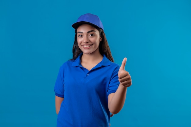Jovem entregadora de uniforme azul e boné olhando para a câmera, sorrindo amigável, feliz e positiva, mostrando os polegares em pé sobre um fundo azul