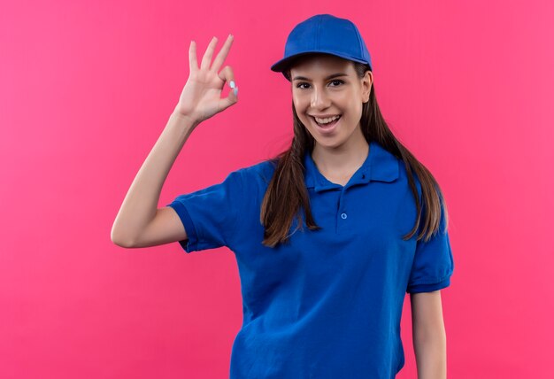 Jovem entregadora de uniforme azul e boné olhando para a camra sorrindo alegremente mostrando sinal de ok