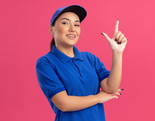 Jovem entregadora de uniforme azul e boné olhando para a frente, feliz e positiva, mostrando o dedo indicador em pé sobre a parede rosa