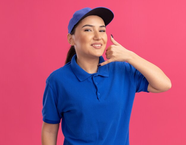 Jovem entregadora de uniforme azul e boné olhando para a frente sorrindo alegremente fazendo um gesto de me chamar de pé sobre a parede rosa
