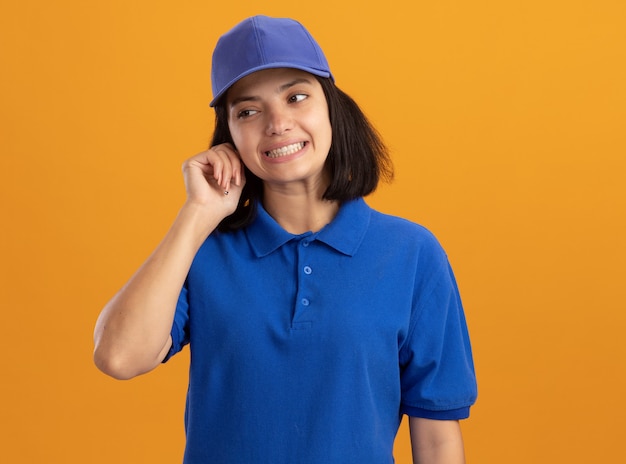 Jovem entregadora de uniforme azul e boné olhando para o lado confusa em pé sobre uma parede laranja