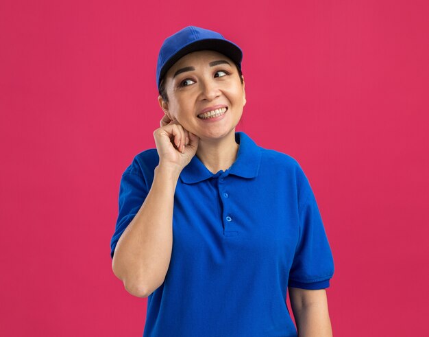 Jovem entregadora de uniforme azul e boné olhando para o lado sorrindo confiante