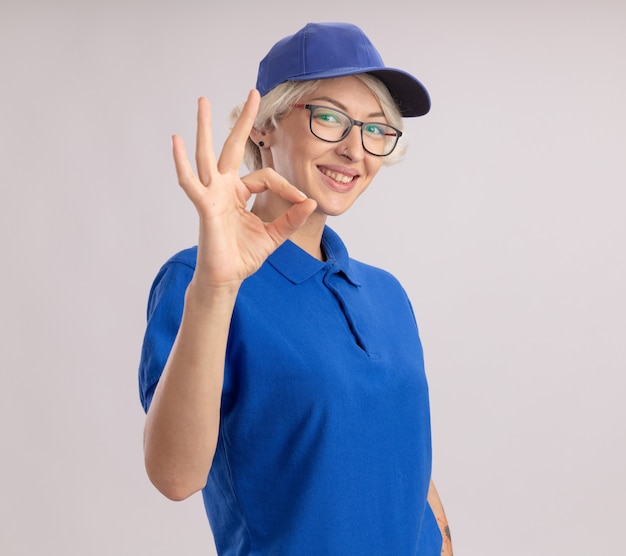 Jovem entregadora de uniforme azul e boné olhando sorrindo mostrando uma placa de ok em pé sobre uma parede branca