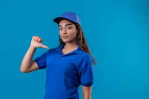 Foto grátis jovem entregadora de uniforme azul e boné parecendo confiante apontando para si mesma com o polegar satisfeita e orgulhosa de pé sobre um fundo azul